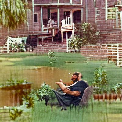 Lean, bearded, older Hispanic man reading a  book, and smoking a tobacco pipe.
He is sitting in a rocking chair on his p...