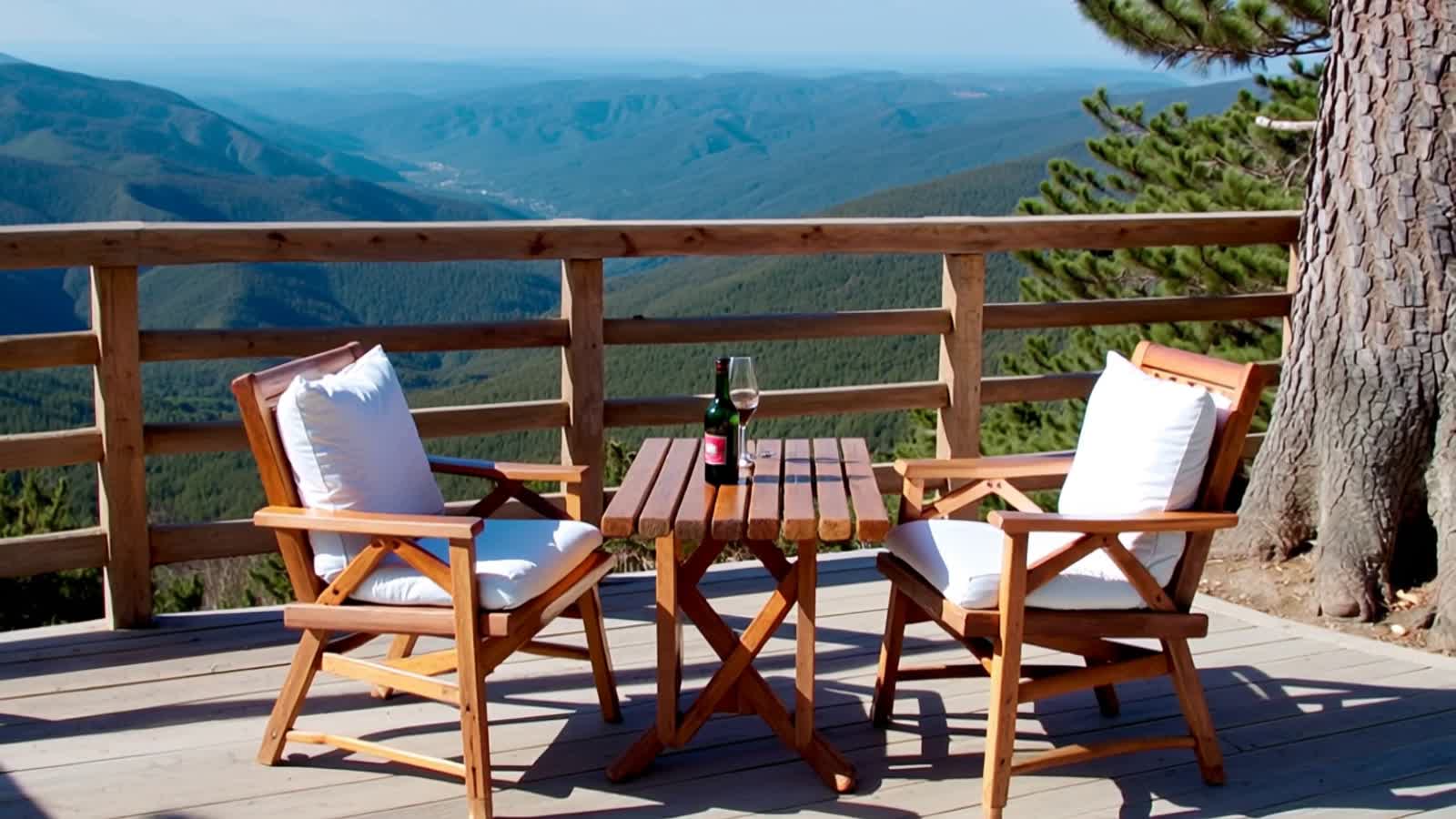 A serene outdoor terrace scene with a panoramic mountain view. In the foreground, a wooden slatted table with a bottle o...