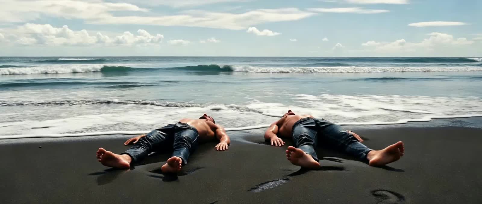 2 Men on a Beach Laying on a Black Sand Beach as the Waves lap gently on their Feet, they're only wearing wet jeans