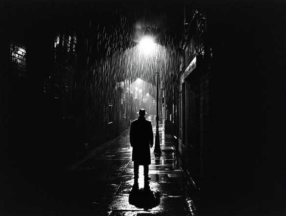 A solitary figure stands under a flickering streetlamp, back turned, in a rain-slicked alley. High-contrast black and white, classic film noir aesthetic, dramatic shadows, gritty texture.