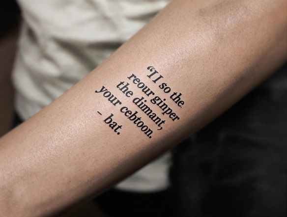 A close-up photograph of a forearm tattoo featuring a quote in a classic typewriter font. The background is blurred, emphasizing the intricate details of the lettering. The font should appear slightly faded, mimicking real ink.