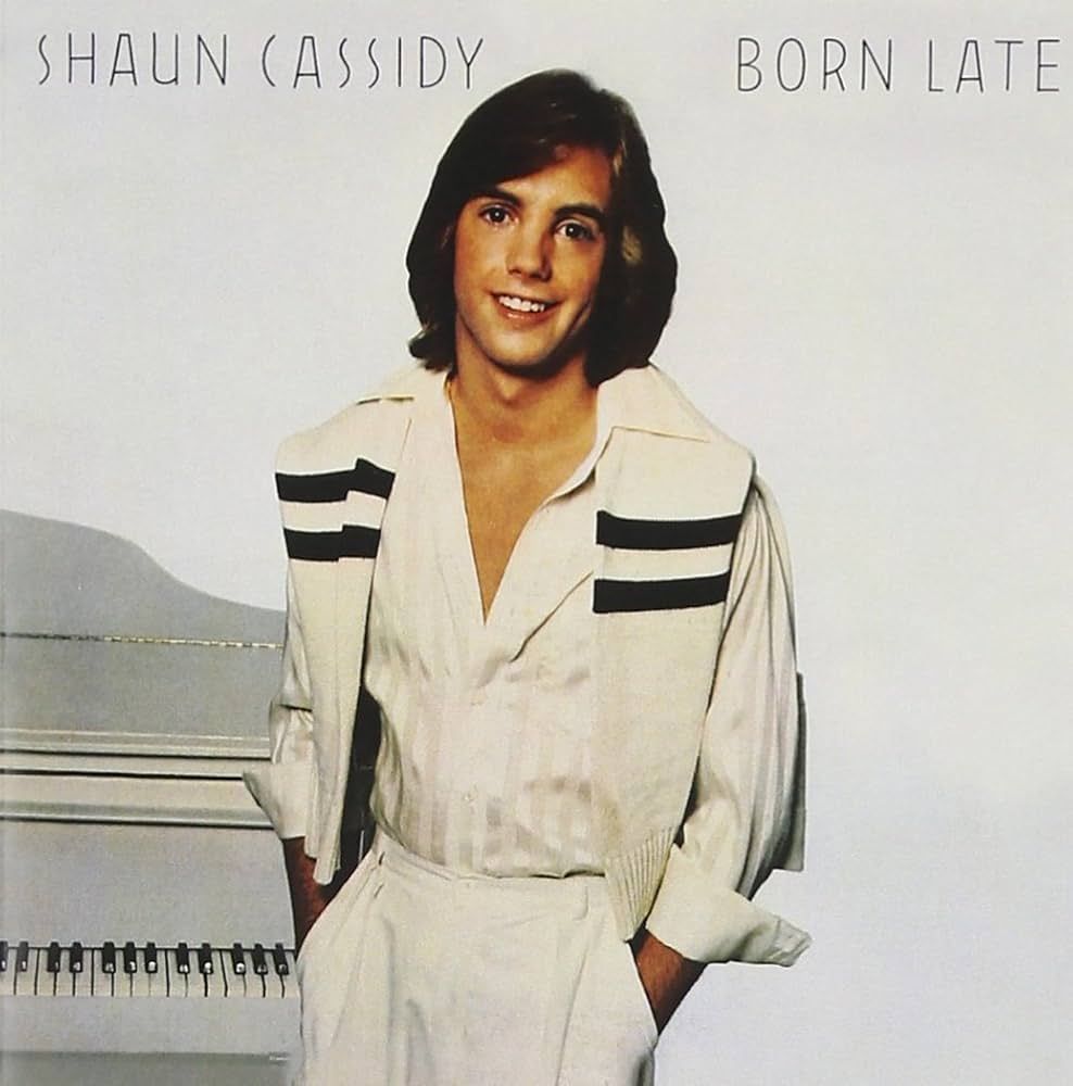Shaun Cassidy stands in front of a glossy white upright piano, dressed in a crisp white outfit with a sleeveless vest featuring bold black v...