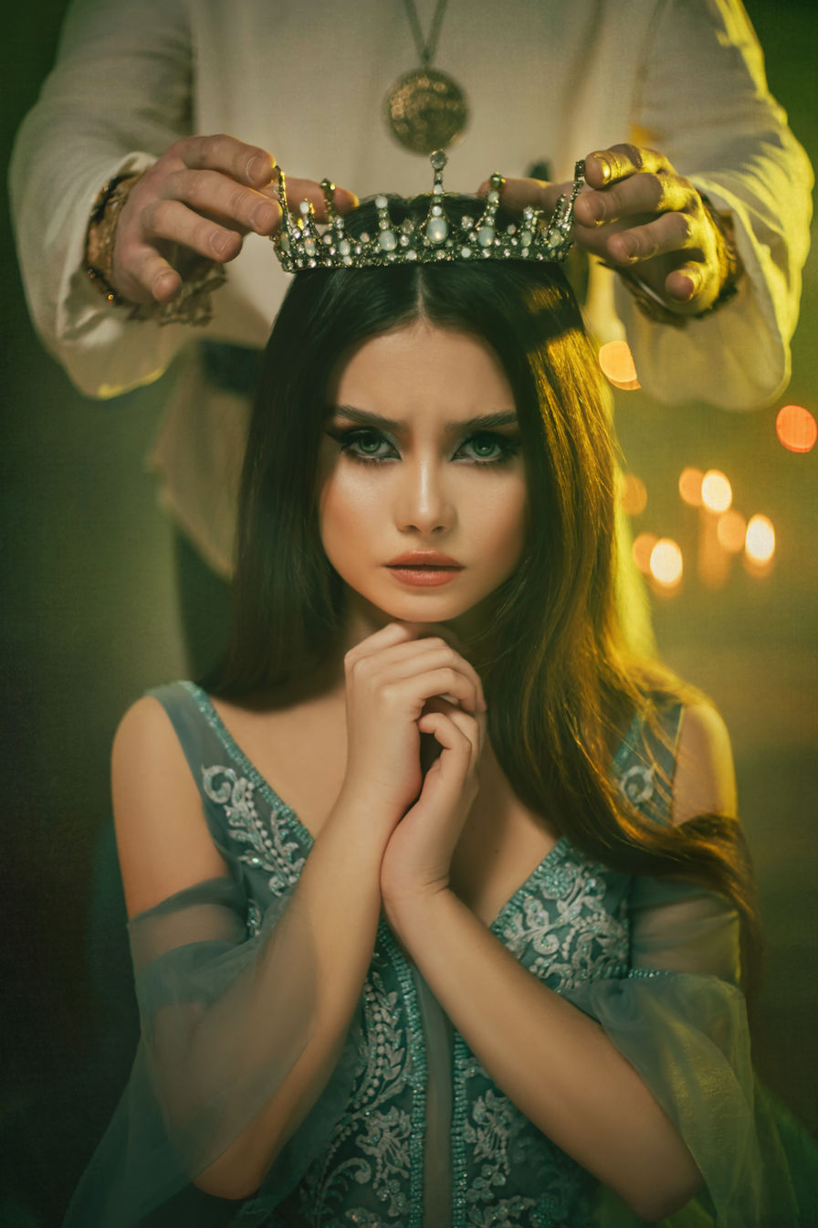 A beautiful black-haired woman, dressed in a white gown with gold embroidery and a gold crown, kneeling and weeping with her hands resting i...