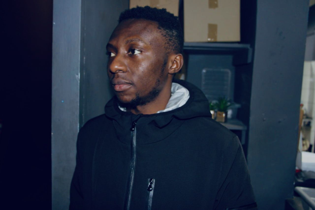 A hyperrealistic portrait of a young African man with glasses, a black cap, and a hoodie, standing in a smoky urban envi...