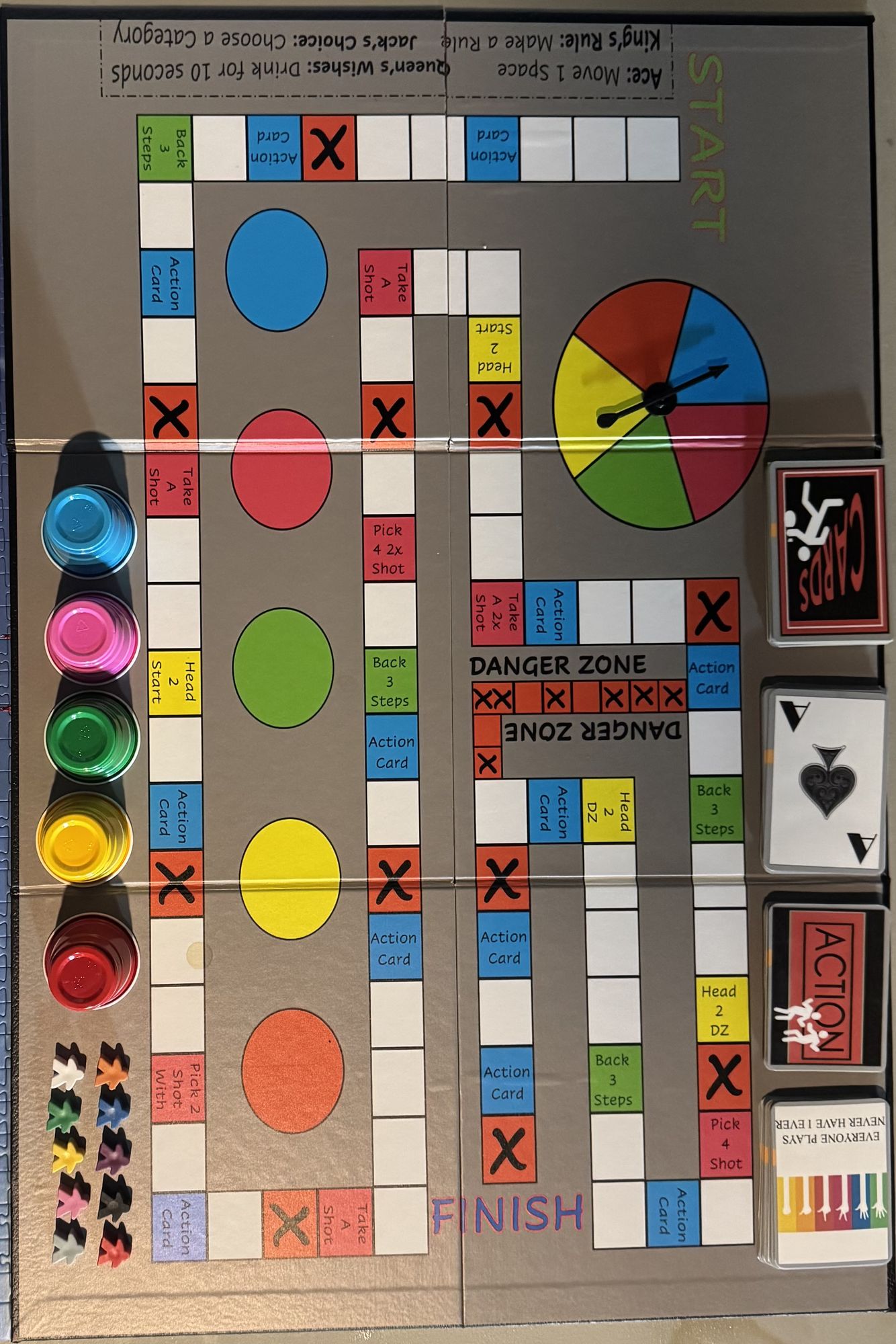 Overhead shot of a beautifully designed board game for 21+ with 10 meeple pieces, roulette spinner, deck of cards and action cards around th...