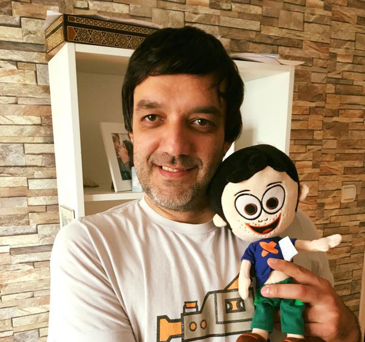 A man with long hair and a short beard is holding a laundry detergent box above his head. The box is blue and says "Merix" on it. He is wear...