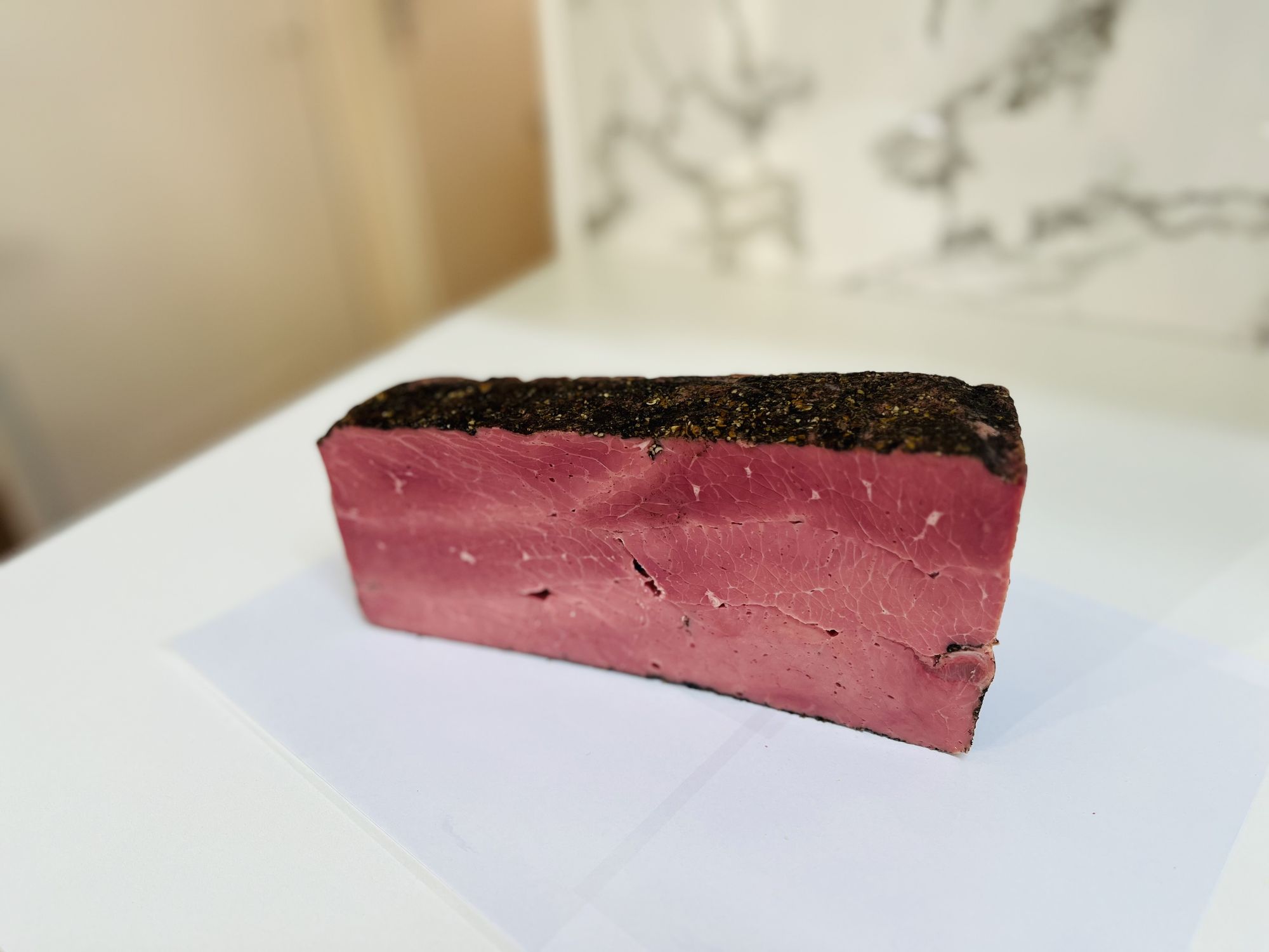 Show this piece of pastrami meat on a wooden plate with some slices in front. With some rosemary ande pickels on the side and black pepper o...