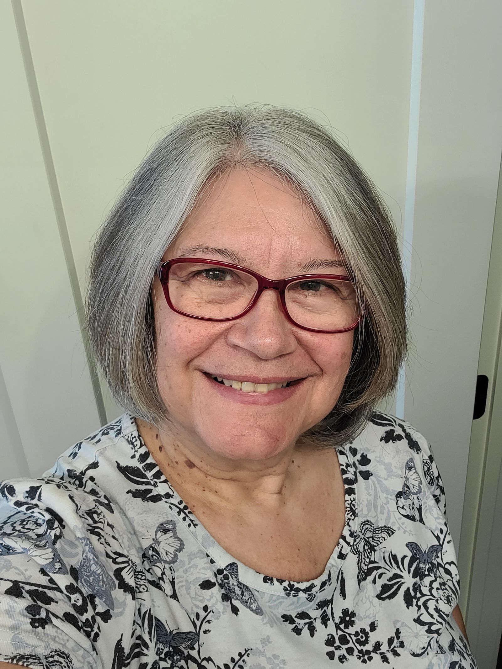 Confident silver-haired woman in her 60s with a warm, witty smile and glasses. Casual-chic style in orange and teal, wor...