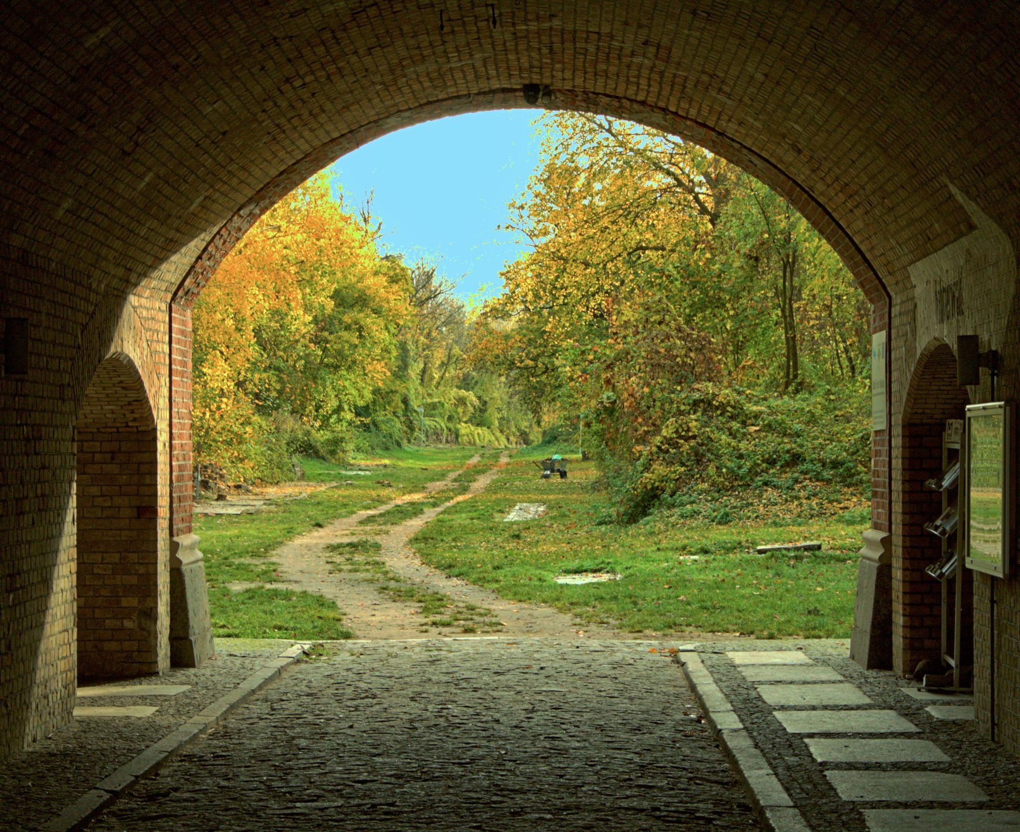 Tall, massive man, he is completely bald, he wears long black coat, he stands before gate, foreground is noir, melancholic, backround seeing...