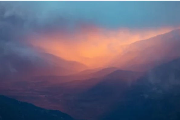 A breathtaking mountain landscape at sunset, with a dramatic interplay of warm and cool colors. Soft layers of mist and clouds drift between...