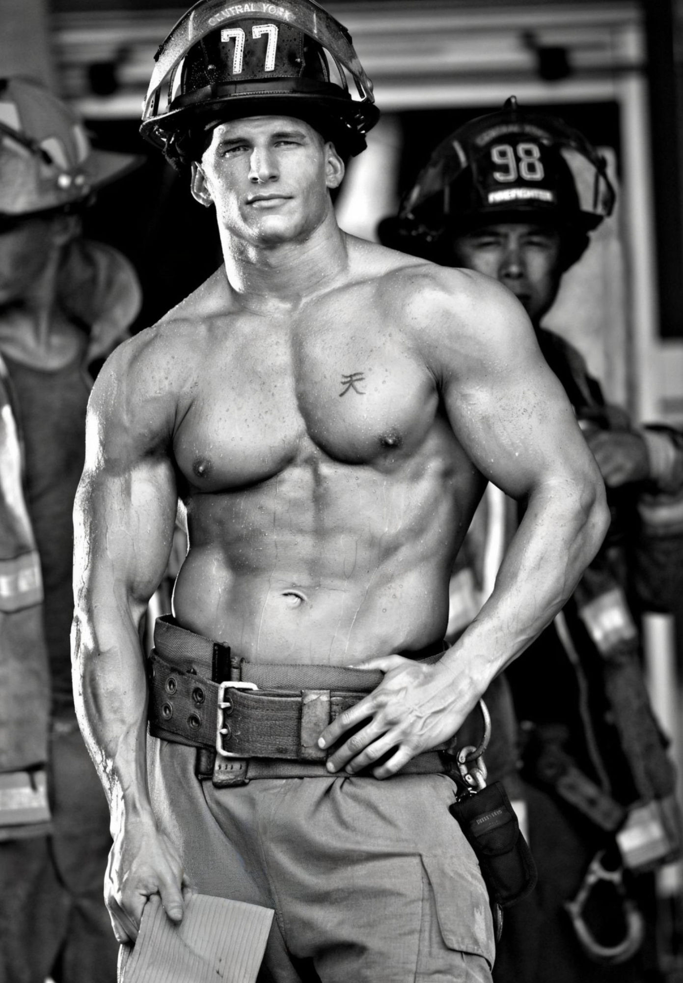 A muscular firefighter stands centered in the frame, his bare chest and defined abs prominently displayed. He wears a yellow and black firef...