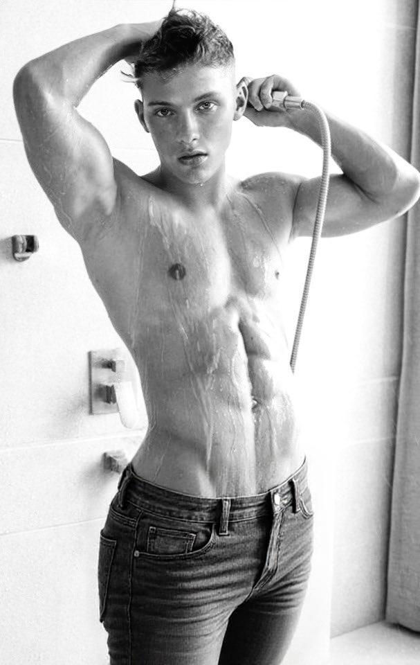 A young, muscular man stands in a shower, facing the viewer with a slight tilt. He is holding a showerhead to his head with one hand, the ho...