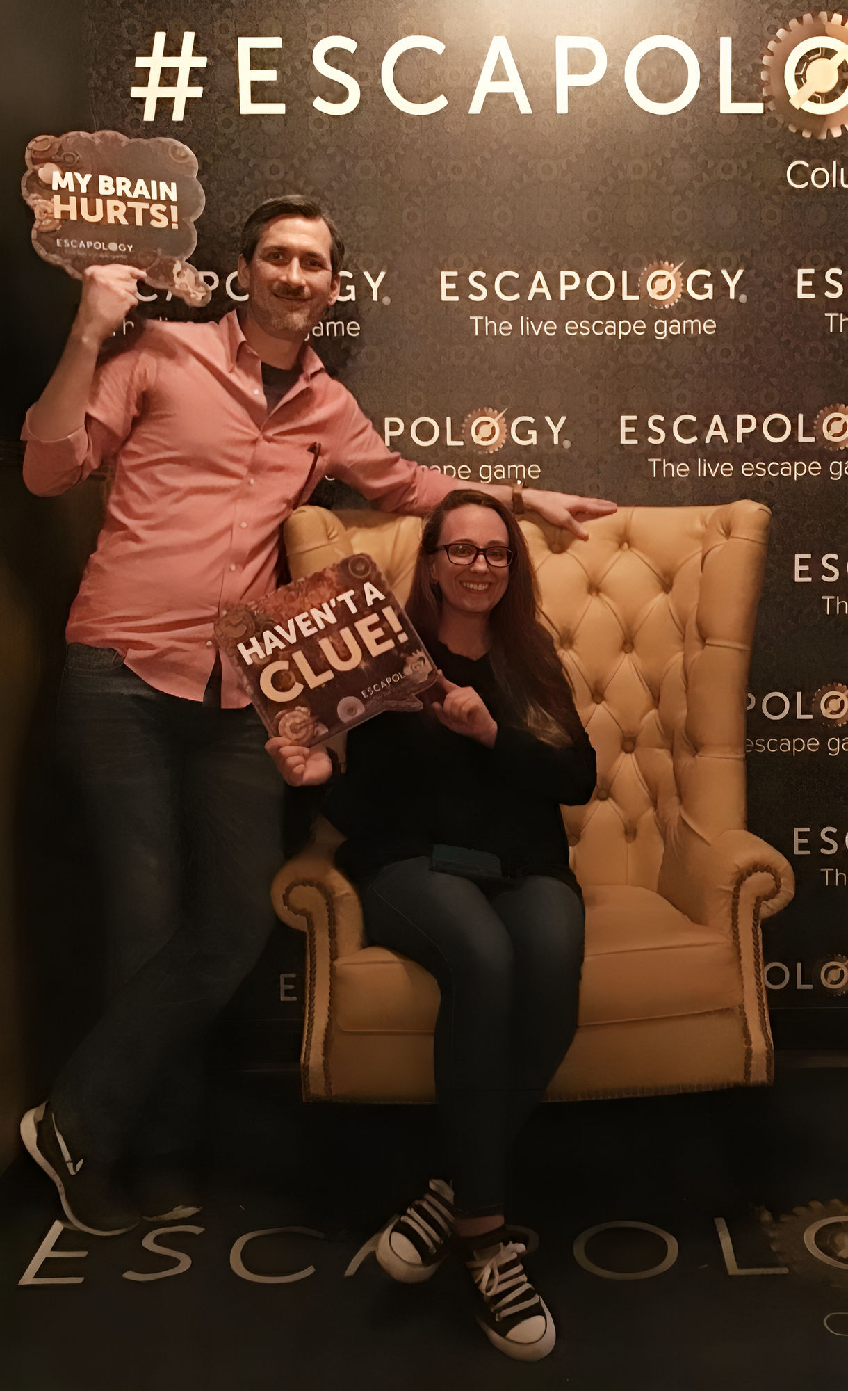 A tall, lean man stands confidently to the left of a yellow vintage armchair with rounded, tufted backrests. He wears a salmon-colored butto...