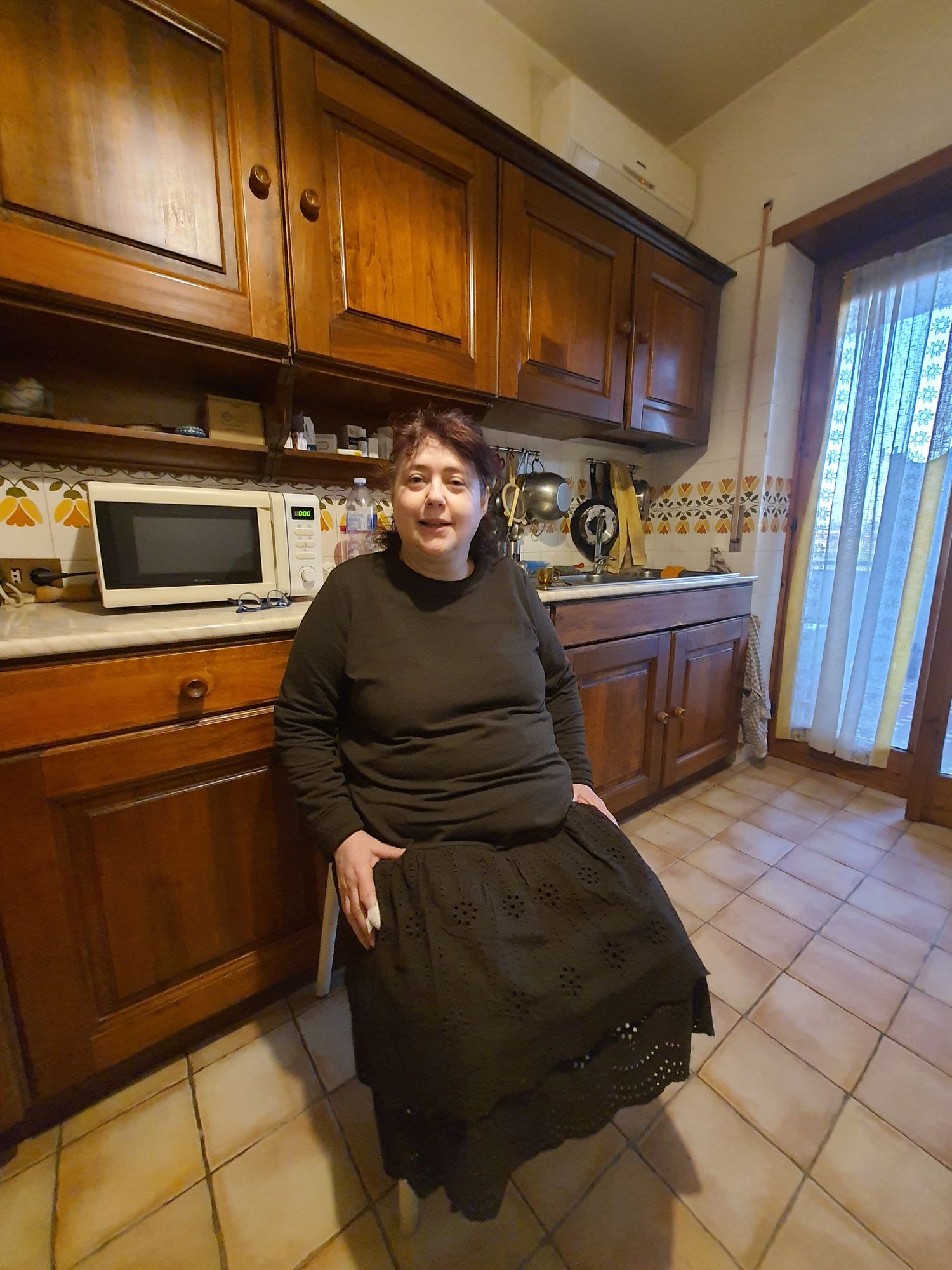 Brunette princess with the hair tied up, in a sangallo brown gown,sitting on a bench in mahogany room. Blue flowers in pots. Window.