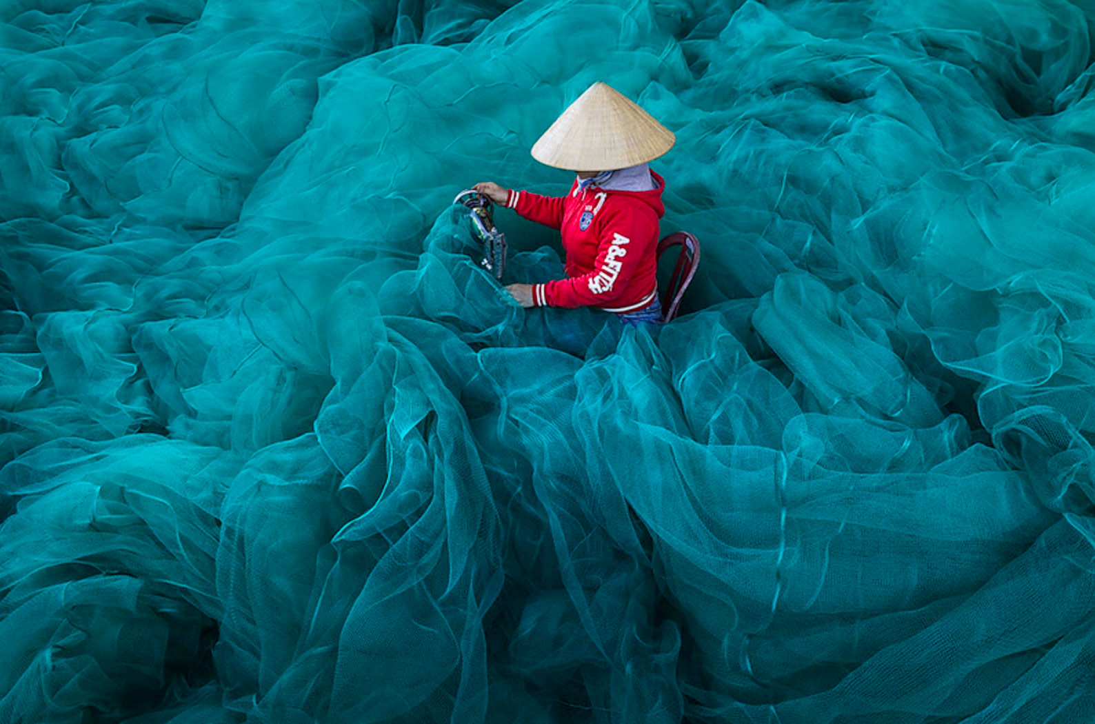 A Vietnamese woman wearing a conical straw hat and a traditional áo dài tunic, mending blue fishing nets with a silver sewing machine in cen...