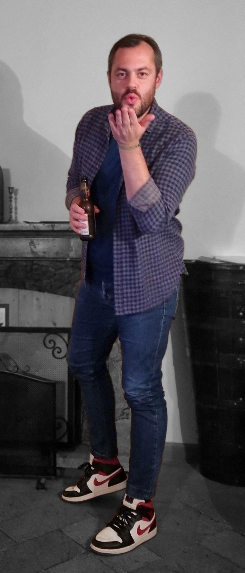  A realistic night-time photo of the same man in the reference image: a bearded man wearing a checkered shirt, dark blue t-shirt, slim jeans...