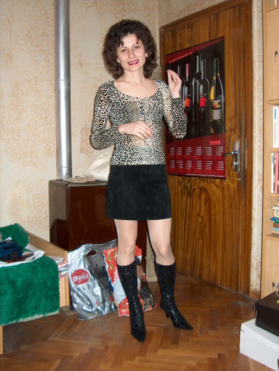 Let the woman be in an elegant clothing boutique. She is standing in front of shelves with beautiful evening dresses. Her hand touches the d...