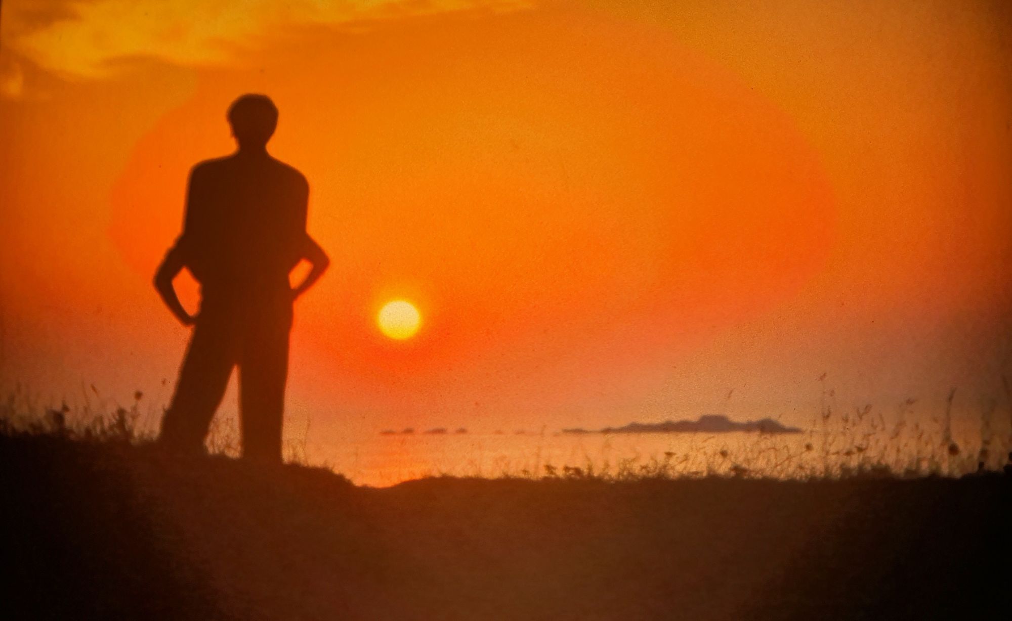 A silhouette of a man stands confidently atop a grassy knoll, hands resting on hips, against a vibrant sunset sky. The man appears to be of ...