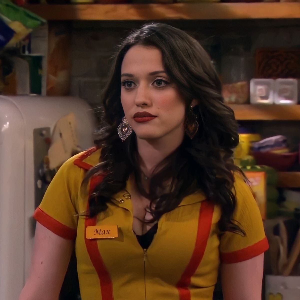 Kat Dennings with long dark hair, wearing a yellow and red uniform with a name tag that reads "Max", looks directly at the viewer with a sli...