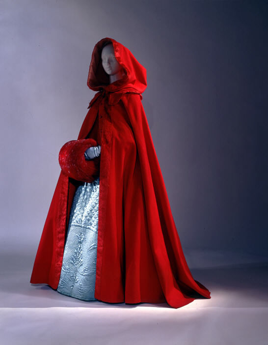 A woman in a deep red hooded cloak, styled like an 18th-century garment with a tied ribbon and ornate clasp at the neck. The hood covers her... - Start Image 2