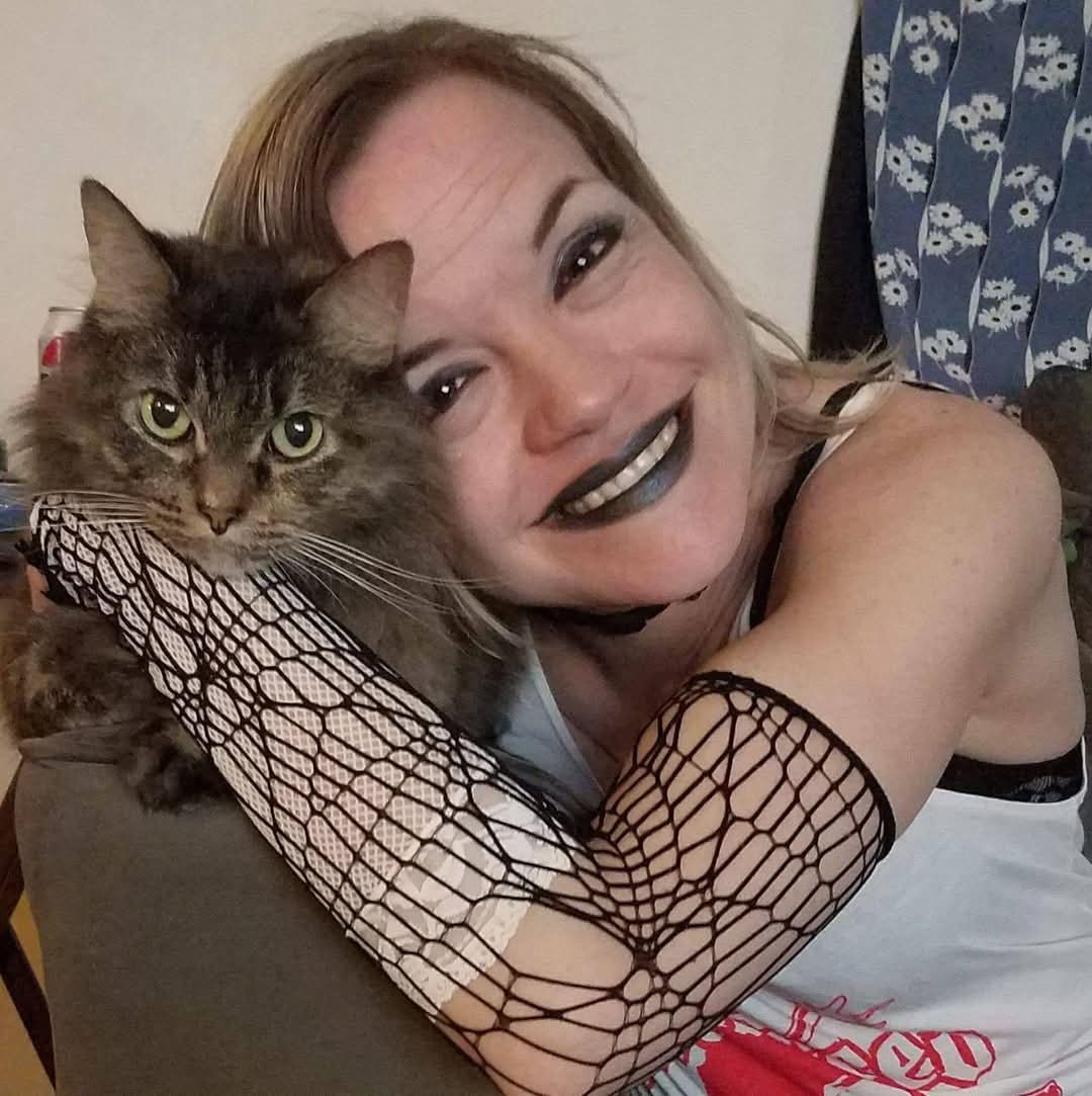 A pretty smiling woman with blonde hair and black lipsticked lips wearing lack fishnet gloves is hugging a beautiful long haired Grey and br...